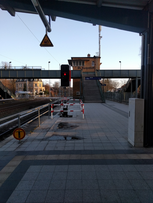 Ein belebter Bahnhof mit einem Zug auf den Schienen, ein Bahnsteig mit Geländern, ein Schild, eine Verkehrsampel an einem Mast, eine Brücke mit Treppen, Laternen, Bäume, Gebäude mit Fenstern, ein entfernter Turm und ein Himmel als Hintergrund.