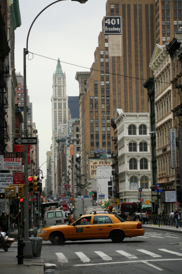 Ein gelbes Taxi fährt auf einer belebten Stadtstraße mit hohen Gebäuden, Fußgängern, Ampeln und Texttafeln, unter einem sichtbaren Himmel.