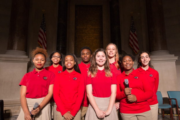 Eine Gruppe junger Frauen in roten Shirts, die strahlen und Mikrofone halten, vor Stühlen, Säulen, Fahnen und einer Wand stehen und damit Teil eines nationalen Jugendchors sind.