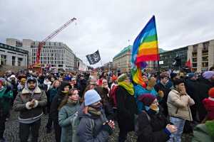 Große Gruppe von Menschen bei der LGBTQ+ Demonstrationen in Berlin, die Fahnen und Plakate halten, mit Gebäuden, einem Kran und einem bewölkten Himmel im Hintergrund.