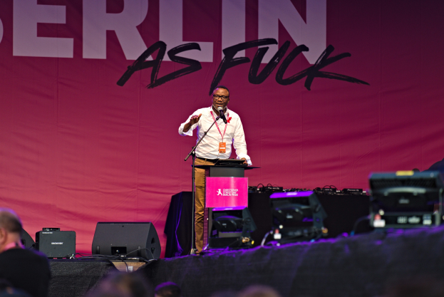 Ein Mann in einem weißen Hemd und braunen Hosen steht auf einer Bühne und hält ein Mikrofon in der Hand, während er zu einer Menge spricht. Im Hintergrund ist ein pinkfarbenes Banner mit der Aufschrift "Berlin as Fuck" zu sehen.