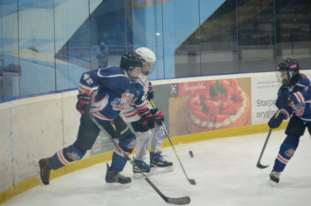Gruppe junger Menschen, die Eisockey auf einer Indoor-Eisfläche spielen, mit Helmen, Sportuniformen und Schlittschuhen sowie Hockey-Schlägern in der Hand, mit einem Plakat im Hintergrund auf einer Glaswand.
