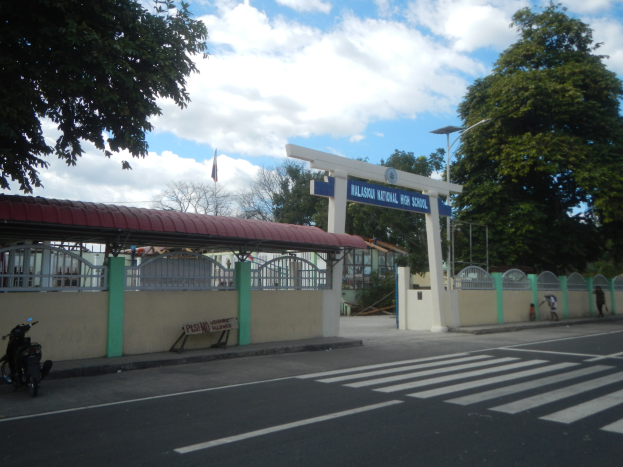 Einfahrt zu einer Schule mit einem vor der Tür geparkten Motorrad, einer Straße mit weißen Linien und Zebrastreifen, einer Mauer mit einem Tor und einem Schuppen, einer Namenstafel, Laternen, Bäumen, einer Fahne auf einem Mast und einem bewölkten Himmel.