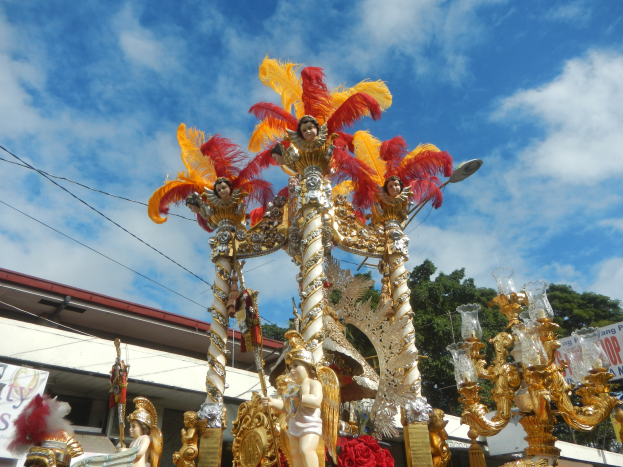 Ein Karnevalswagen, geschmückt mit goldenen und roten Federn, der Menschen trägt, wird in einem Umzug mit einem Gebäude, Bäumen, Drähten und einem Banner in der Nähe unter einem sichtbaren Himmel gezeigt.