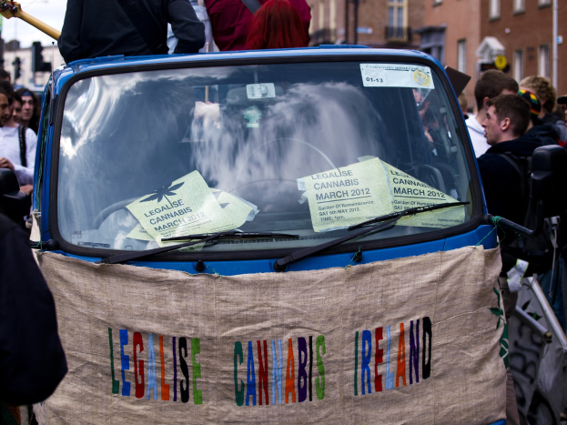 Gruppe von Menschen, die um ein Auto mit einem "Legalise Cannabis Ireland"-Schild stehen, umgeben von Gebäuden unter einem klaren blauen Himmel, mit Papieren im Auto sichtbar.