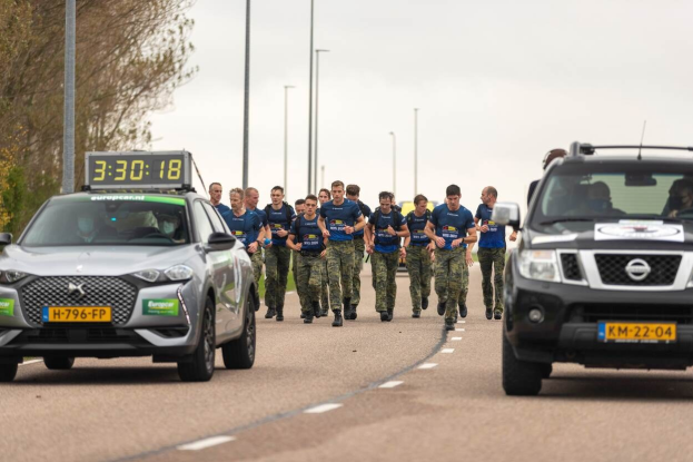 Eine Gruppe von Menschen, die auf einer Straße neben parkenden Autos gehen, mit Bäumen auf der linken Seite und Strommasten im Hintergrund während eines Outdoor-Events.