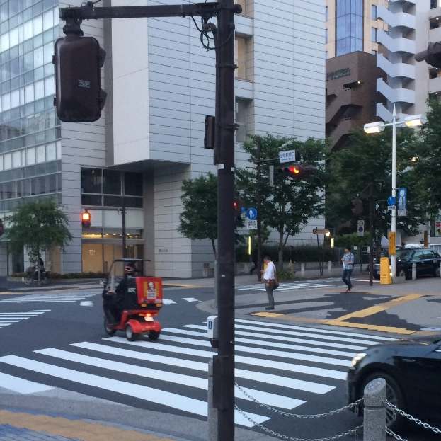 E-Scooter auf einer Straße mit Passanten im Hintergrund, einer Ampel im Vordergrund und Gebäuden mit Bäumen dahinter.