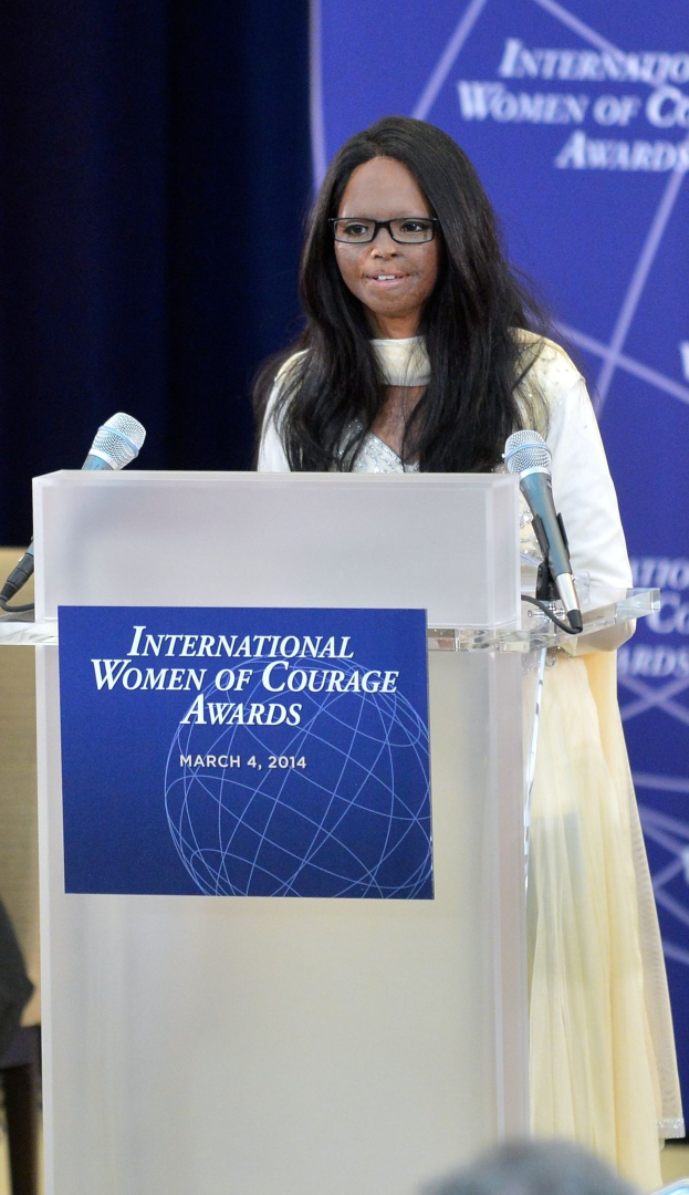 Frau in weißem Kleid und Brille spricht an einem Pult mit zwei Mikrofonen und einer Tafel während der International Women of Courage Awards 2014.