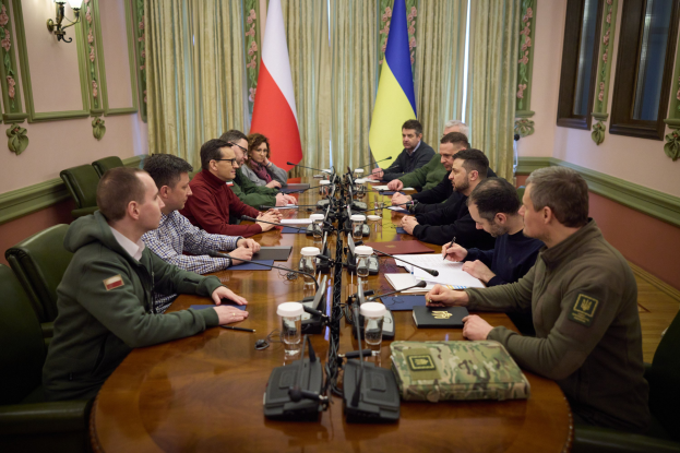 Gruppe von Männern in formeller Kleidung, die um einen Holztisch mit Mikrofonen und Dokumenten sitzen und in einem Raum mit Flaggen und an der Wand montierten Lampen diskutieren.