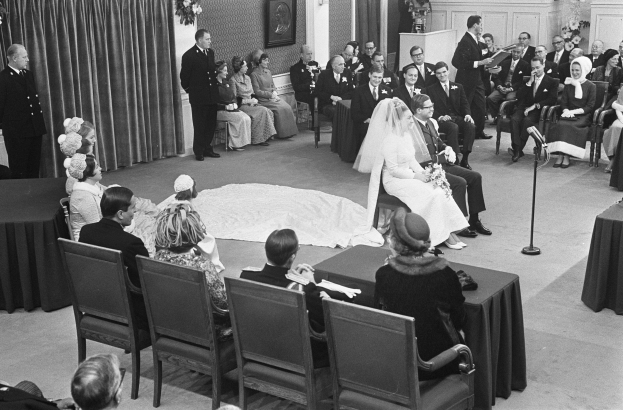 Schwarz-weiß-Foto einer Hochzeitszeremonie mit sitzenden und stehenden Menschen, einem Mann mit Mikrofon und einer Frau mit Blumenstrauß, sowie einem Rednerpult, Blumenschmuck, einem Vorhang, einem Rahmen und einer Tür im Hintergrund.