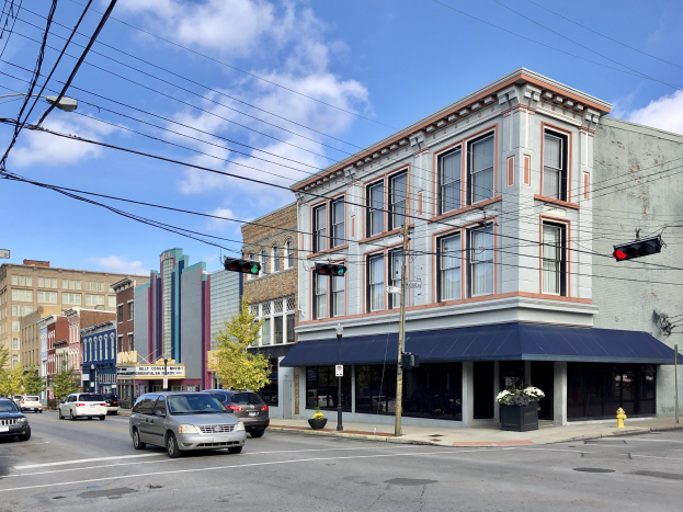 Eine Straße in einer Stadt mit geparkten Autos, Strommästen, Ampeln, Straßenlaternen, Bäumen und Gebäuden, unter einem bewölktem Himmel, mit Text, der ein kommerzielles Grundstück zum Verkauf für $1,250,000 anzeigt.