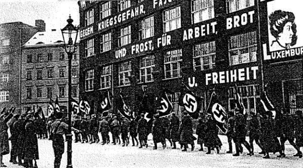 Schwarzes und weißes Bild einer Gruppe, die mit Fahnen und Transparenten die Straße entlangmarschiert, mit einem Lichtmast links und einem Gebäude mit einem Nazi-Parteischild im Hintergrund.