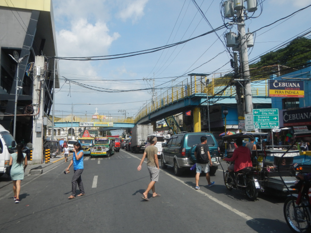 Eine belebte Stadtstraße mit Autos, Motorrädern, Fußgängern, Gebäuden, Strommasten, Schildern, einer Brücke, Bäumen und einem bewölkten Himmel.