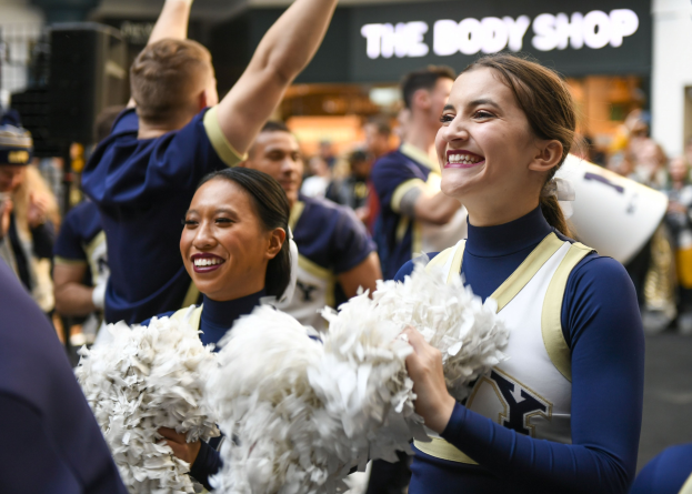 Eine Gruppe von Cheerleadern der University of West Virginia in blauen und weißen Uniformen, die vor einem Publikum lächeln und Pompons halten, mit einem Gebäude im Hintergrund, das "University of West Virginia" bezeichnet ist.