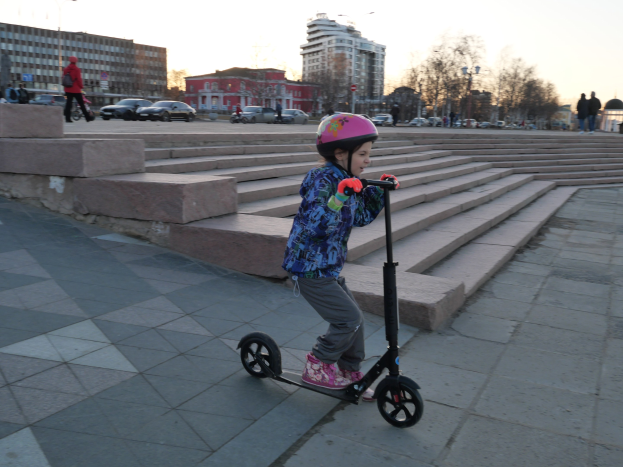 Ein junges Mädchen in einem Helm fährt auf einem Scooter eine Treppe hinunter mit Fahrzeugen, Menschen, Laternenmästen, Bäumen, Gebäuden und einem klaren blauen Himmel im Hintergrund.