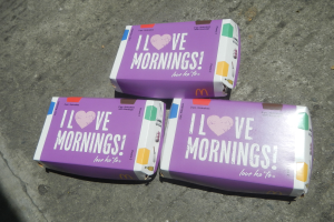 Drei buntfarbene McDonald's 'Ich liebe morgens'-Schachteln mit dem McDonald's-Logo und fetter weißer Schrift auf einem hellblauen Himmel mit weißen Wolken.