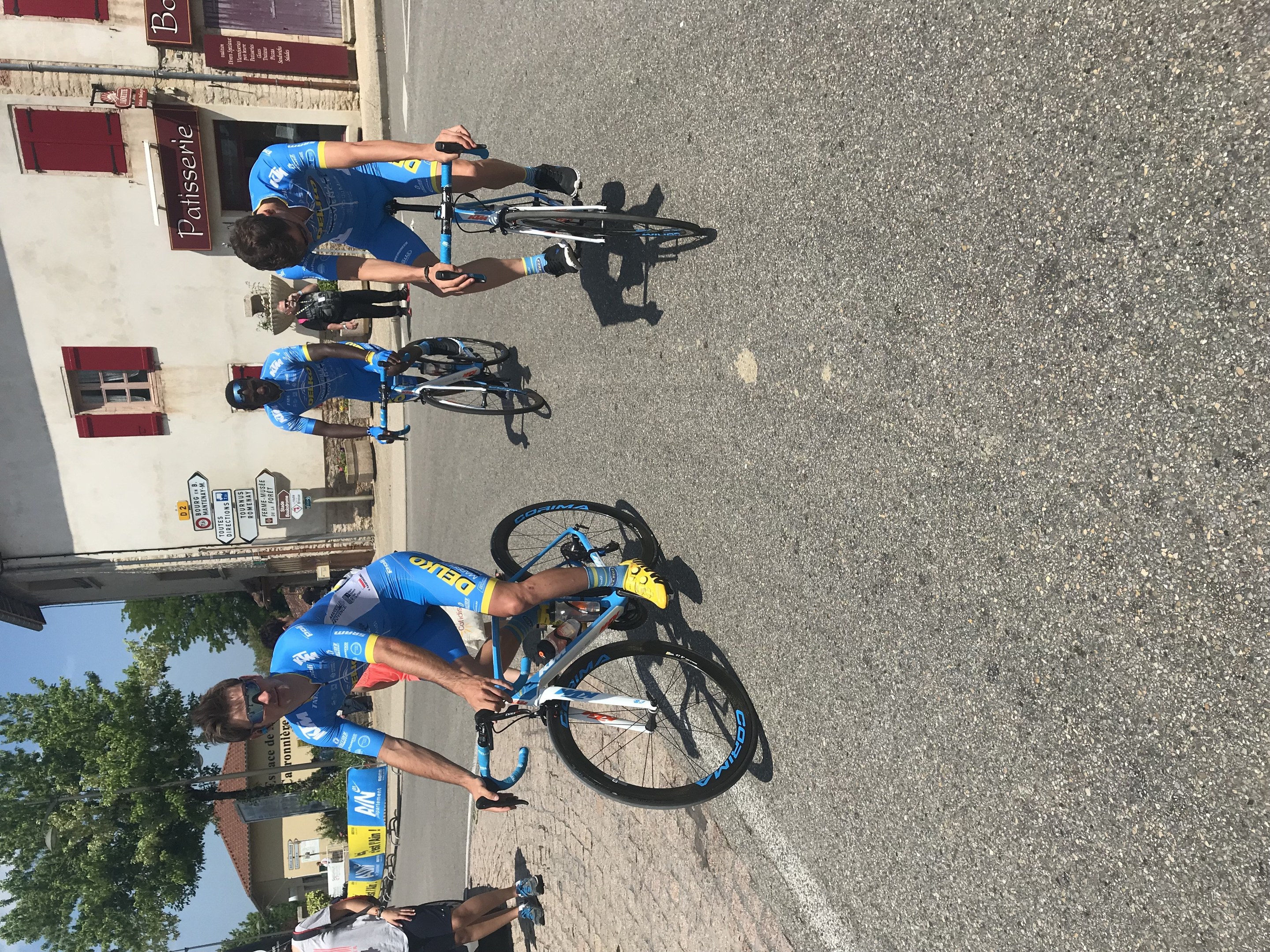 Eine Gruppe von Radfahrern fährt eine Straße entlang an einem Gebäude vorbei, mit Fenstern und Schildern, Bäumen und einem klaren Himmel im Hintergrund und Zuschauern auf der Straße darunter.