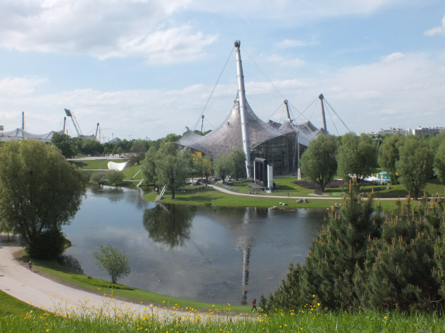 Olympiastadion in London, England, umgeben von Grünflächen und Blumen, mit einer Straße, einem Gewässer, Gebäuden und einem bewölkten Himmel im Hintergrund.
