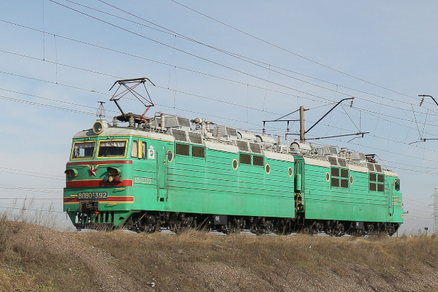 Ein grüner Elektrolokomotive fährt auf Schienen durch ein grünes Feld mit Strommasten und einer fernen Turm unter einem klaren blauen Himmel.