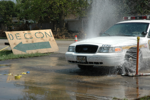 Ein Streifenwagen fährt durch eine Pfütze, mit einem Schild, einer Verkehrskegel und anderen Gegenständen am Boden, vor einem Hintergrund aus Bäumen, Gebäuden und einem klaren blauen Himmel.