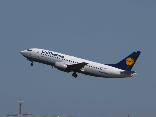 Lufthansa Airbus A320-200 im Flug vor einem klaren blauen Himmel mit Gebäuden und Polen im Hintergrund.