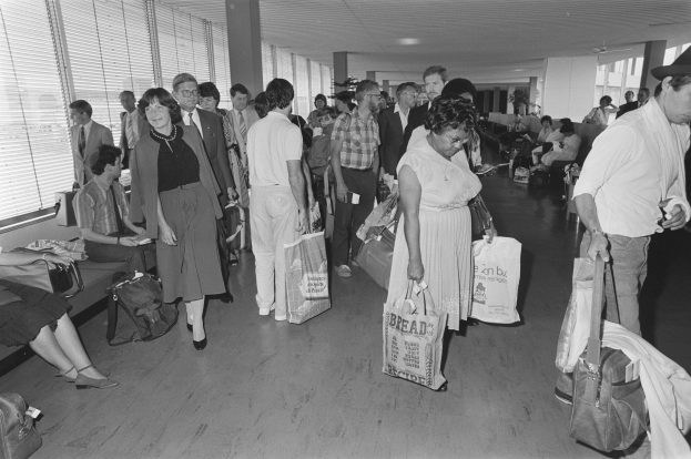Schwarzes und weißes Bild von Reisenden in einem Flughafen, einige sitzen auf Bänken und andere stehen mit Gepäck, mit einem Mann und einer Frau, die Taschen halten, in der Mitte und Fensterjalousien auf der linken Seite.