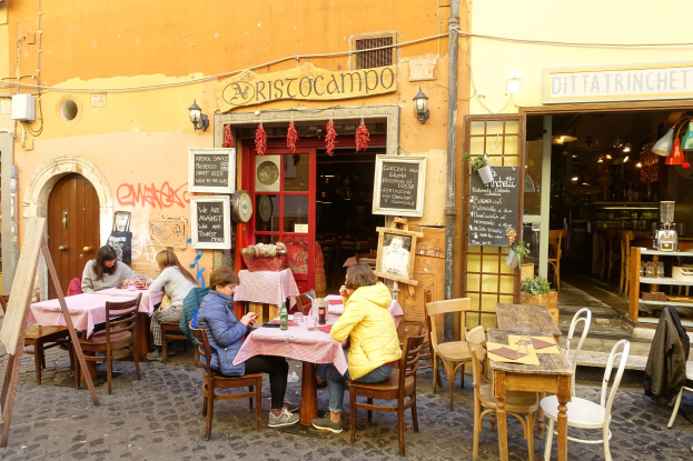 Gruppe von Menschen an Tischen eines Restaurants in Rom, Italien, mit Flaschen, Gläsern und beleuchteter Dekoration, einschließlich Texttafeln an den Wänden und Regalen mit Gegenständen im Inneren.