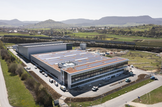 Luftbild eines großen Gebäudes mit Solarmodulen auf dem Dach, umgeben von Fahrzeugen, Bäumen, Gras und Strommasten, vor Bergen und einem klaren blauen Himmel; das Gebäude ist Deutschlands erste solarbetriebene Fabrik.