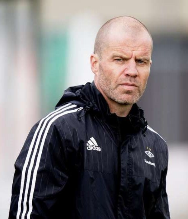 Kahlköpfiger Trainer in marineblauer Jacke mit weißen Streifen und weißem Kragen, der auf einem Fußballfeld steht und nachdenklich die Hände vor der Brust verschränkt.