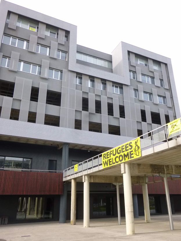 Ein großes Gebäude mit einem "Flüchtlinge Willkommen"-Schild, umgeben von Säulen und einem Geländer, vor einem Himmel-Hintergrund.