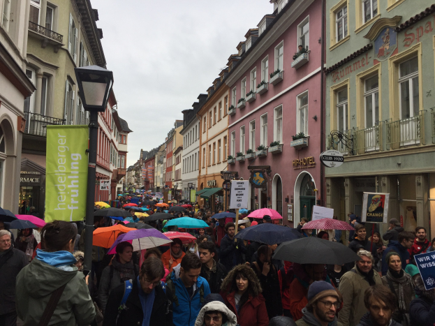 Eine große Gruppe von Menschen geht mit Schirmen die Straße entlang, einige tragen Taschen und halten Schilder, während sie an einem Lichtmast und Gebäuden mit Fenstern vorbeigehen.