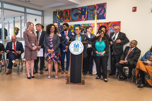 Eine Gruppe von Menschen steht vor einem Podium mit einem Mikrofon und einer Tafel mit der Aufschrift "Sancho-Stadtrat tritt dem Stadtrat bei", mit anderen im Hintergrund sitzend und stehend, Flaggen, einer Tafel, einem Feuerlöscher und Deckenleuchten an der Rückwand.