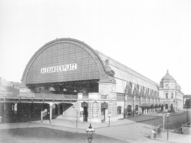Ein Schwarz-Weiß-Foto von Alexanderplatz in Berlin, Deutschland, mit einem großen Gebäude mit Säulen, Bögen und einer Namensplakette, Laternen und Menschen auf der Straße, sowie Bäumen und einem klaren Himmel im Hintergrund.
