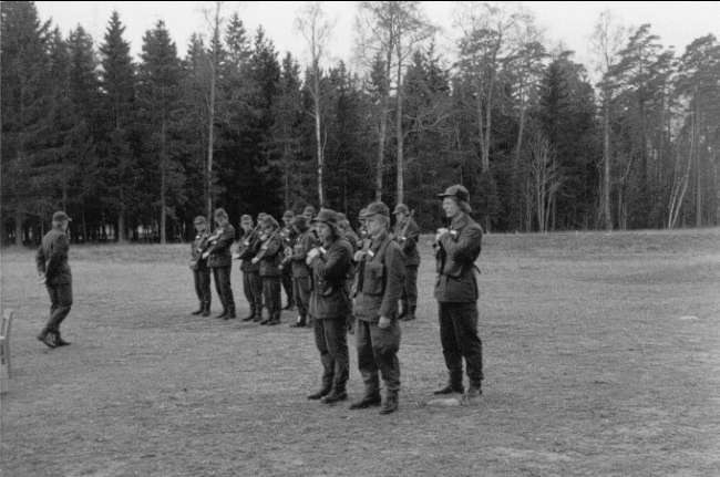 Schwarzes und weißes Bild einer Gruppe von Männern in Mützen mit Gewehren, die in einem Feld mit Bäumen und einem klaren Himmel im Hintergrund stehen.