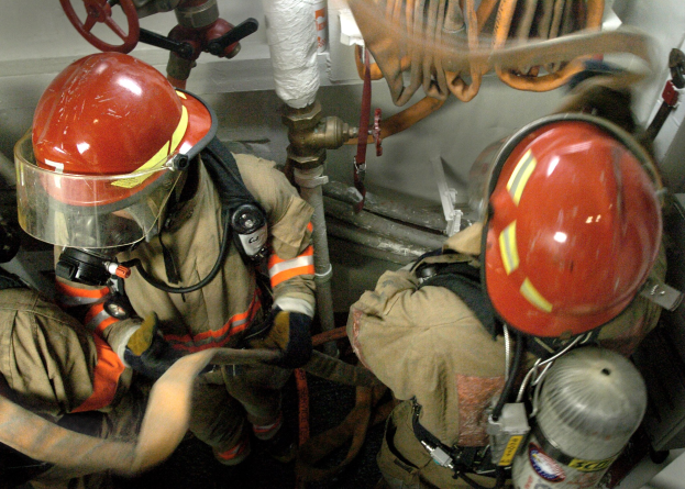 Feuerwehrleute in Helmen und Schutzausrüstung arbeiten an einem Feuerhydranten mit Rohren, Ventilen und einer Wand im Hintergrund.