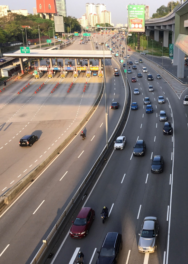 Eine vielbefahrene Autobahn mit zahlreichen Autos und Motorrädern, Verkehrsleitkegeln, Hinweisschildern, Laternenmasten, einer Brücke, Bäumen, Gebäuden und einem klaren blauen Himmel.