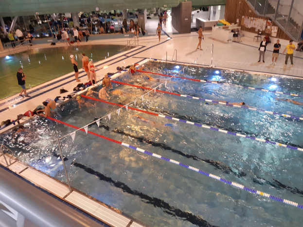 Großes Hallenbad mit schwimmenden Menschen, anderen am Beckenrand und Treppen auf der rechten Seite, mit einer Wand im Hintergrund.