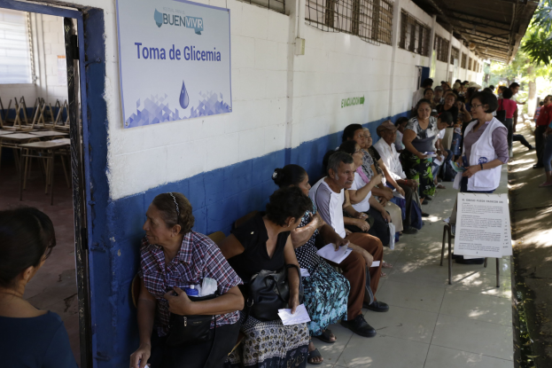 Menschen sitzen auf Bänken vor einem Gebäude an einer Wahllokal in Guadalajara, Mexiko, mit einer Tafel an der Wand, einer Tür links und Bäumen im Hintergrund.
