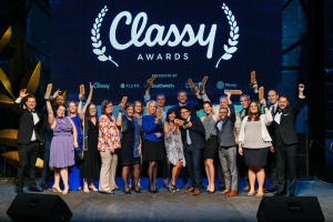 Gruppe von Menschen auf einer Bühne, die Auszeichnungen halten, mit Lautsprechern und einem Bildschirm im Hintergrund, der "Classy Awards 2019" anzeigt.
