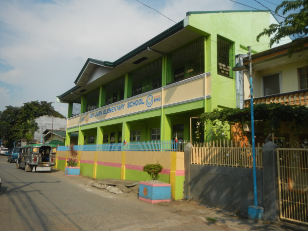 Schulgebäude mit grüner und gelber Fassade, umgeben von Fahrzeugen, Topfpflanzen, einem Zaun, einem Tor, einem Straßenpfosten, Bäumen, Drähten und einem bewölkten Himmel.