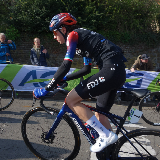 Die professionelle Radrennfahrerin Katie Archibald fährt ein Fahrrad auf einer Straße, trägt einen Helm, eine Schutzbrille, Handschuhe und Schuhe und fährt an einer Menschenmenge vorbei. Im Hintergrund gibt es ein Banner mit Text, eine Wand, Bäume und ein Gebäude.