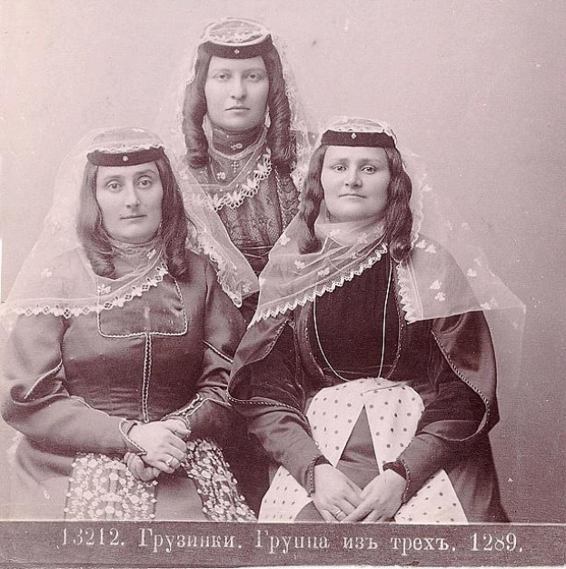 Drei Frauen in traditionellen russischen Hochzeitskleidern mit farbenfroher Stickerei und Perlen, die vor einer weißen Wand in einem Schwarz-Weiß-Foto posieren.