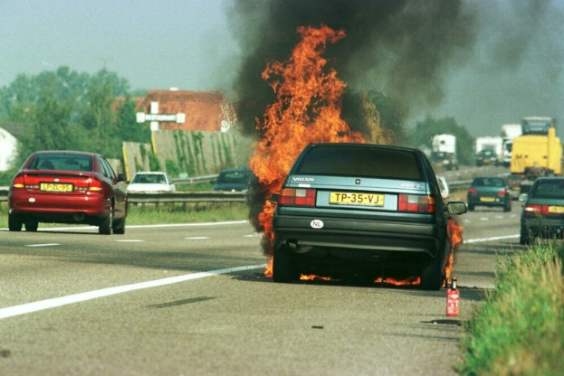Ein Auto, das in Flammen steht, am Straßenrand umgeben von anderen Fahrzeugen, mit Bäumen, Gebäuden und einem klaren blauen Himmel im Hintergrund und einem Feuerlöscher auf der rechten Seite.