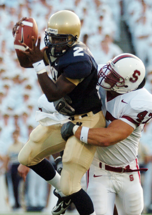 Zwei Fußballspieler in Helmen und Uniformen; einer fängt einen Ball in der Luft, während er von dem anderen getackelt wird.