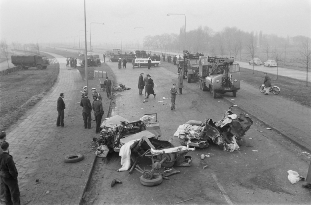 Schwarz-weiß-Szene eines Autounfalls am Straßenrand mit mehreren Fahrzeugen und einer Gruppe von Menschen drumherum, Lichtmasten, Bäume und der Himmel im Hintergrund.