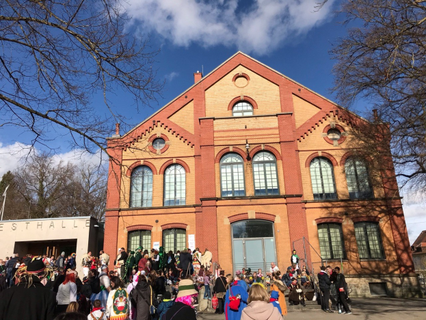 Große Gruppe von Menschen, die vor einem mehrstöckigen Gebäude mit Fenstern und Türen stehen, umgeben von Bäumen unter einem bewölkten Himmel, einige tragen Mützen und tragen Taschen.