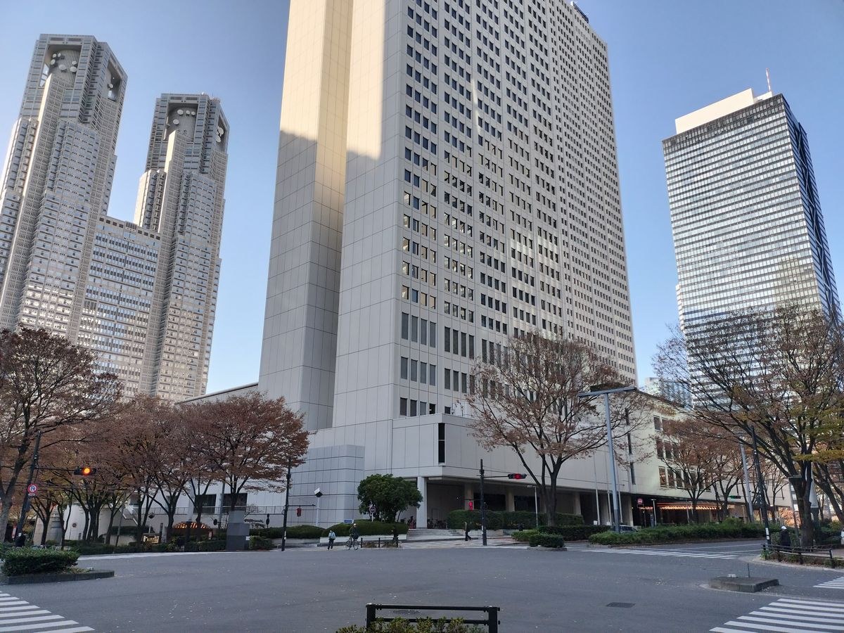 Eine Stadtstraße in Tokio, Japan, mit hohen Gebäuden, Bäumen, Laternen, Verkehrszeichen und einigen Menschen, wobei das Tokyo Metropolitan Government Building im Hintergrund zu sehen ist.