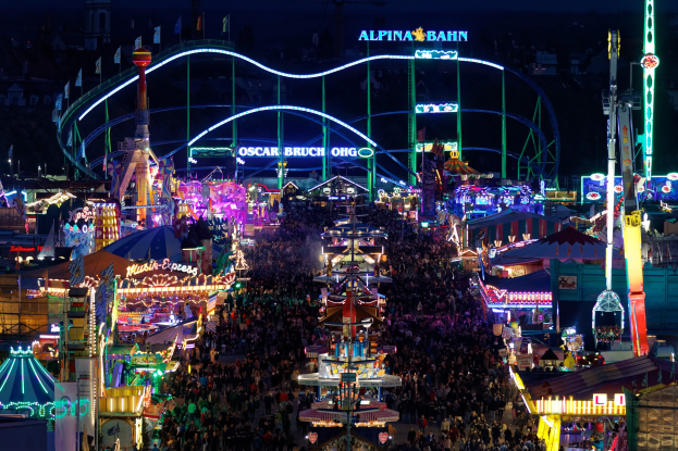 Eine große Menschenmenge bei einem nächtlichen Karneval mit bunten Lichtern, Fahrgeschäften, Pfählen, Fahnen und Namensschildern im Dunkeln.