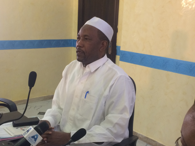 Ein Mann in einem weißen Gewand und Hut sitzt an einem Tisch mit Mikrofonen, spricht, während Papiere und Gegenstände auf dem Tisch sichtbar sind, mit einer anderen Person mit Brille rechts von ihm, in einem Raum mit einer Wand und einer Tür im Hintergrund.