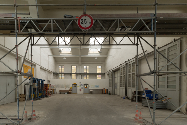 Ein großes Lager mit ausgedehnten Gerüsten, Verkehrskegeln auf dem Boden, Metallstangen, ein Schild, verschiedene Maschinen, eine Tür, Fenster, eine Wand und ein Dach mit Deckenbeleuchtung.